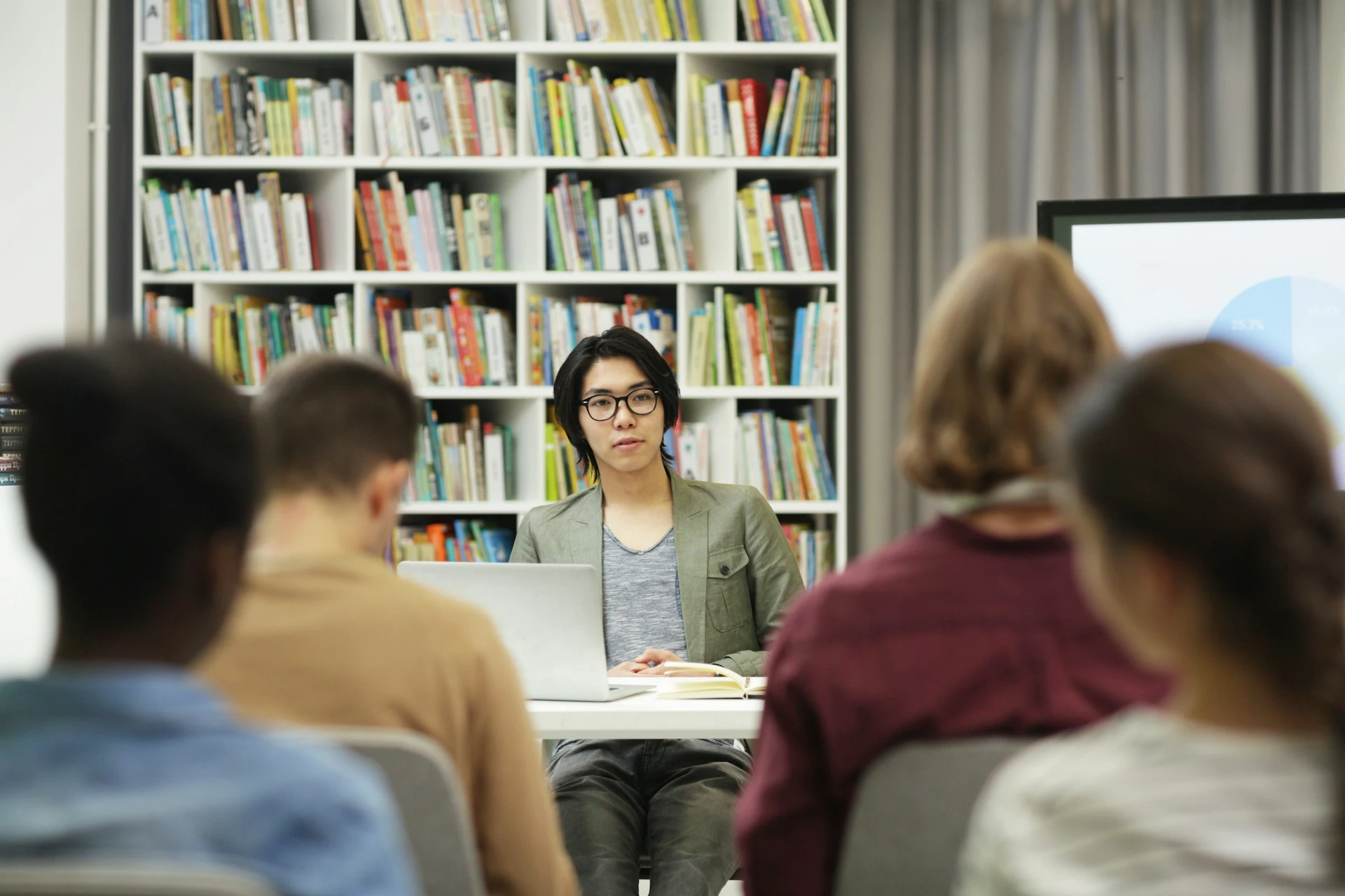 An instructor sitting at a desk addressing a class in front of bookshelves in a library.
